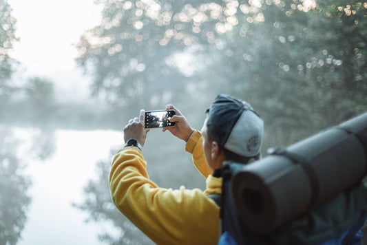 A man in a yellow jacket takes a photo of a misty lake with his smartphone in the morning outdoors.