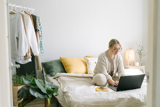 A woman working from home on a laptop in a cozy, stylish bedroom setting.